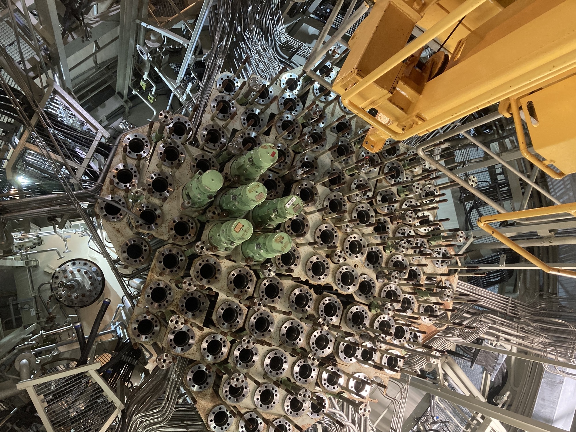 Blick nach oben im Raum unter den Reaktor, es ist eine Art Gitter aus Löchern, in denen sich Brennelemente befinden würden, in ein paar von den Löchern sind die grünen Motoren der Steuerstäbe verbaut, rechts unten ragt eine gelbe Metallplattform nach oben Richtung der Steuerstäbe