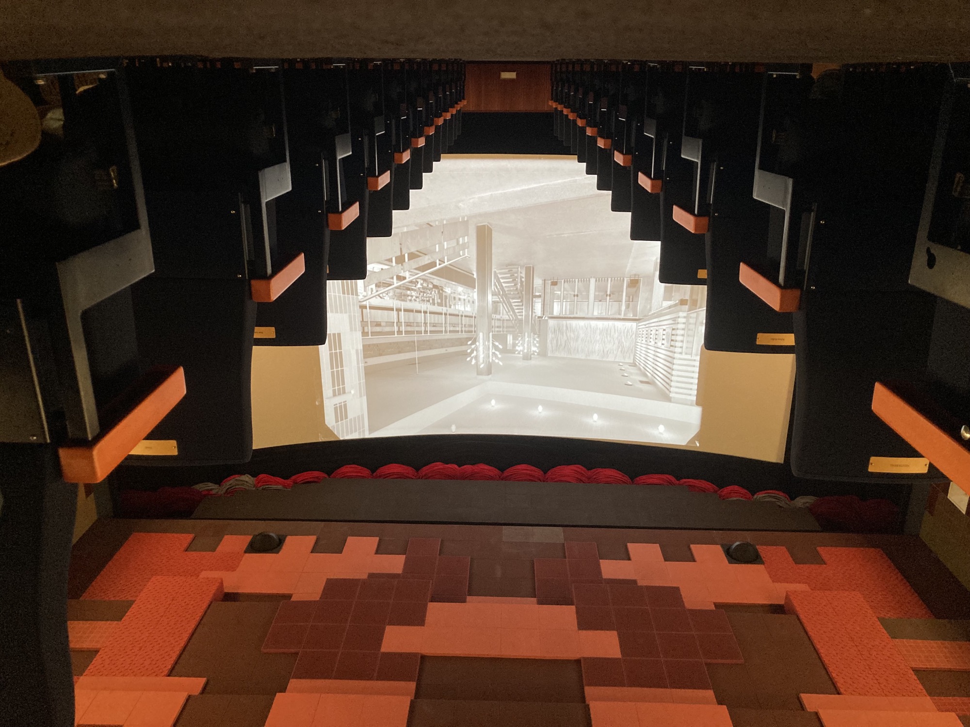 Bodenperspektive mittig im Kinosaal, links und rechts die Sitzreihen mit orangefarbenen Armstützen, mittig die Leinwand, die ein Schwarz-Weiß-Bild des Kinofoyers zeigt, darüber die in rötlichen Tönen gestaltete Decke des Kinosaals