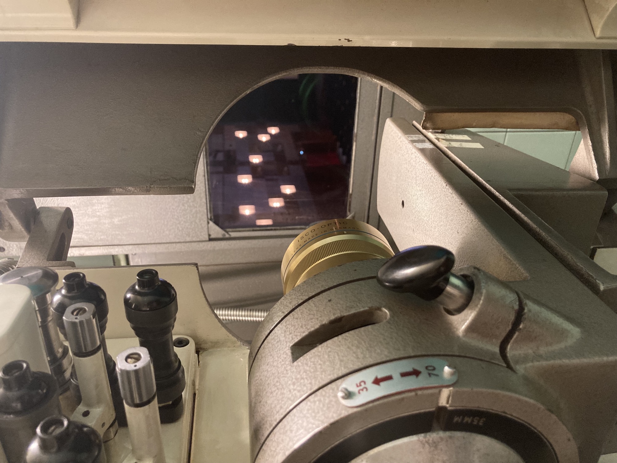 ein Projektor in Blickrichtung auf das Fenster, hinter dem die Kinoleinwand zu sehen ist, im Vordergrund ein Umschalter zwischen 35 und 70, im Mittelgrund das goldene Objektiv