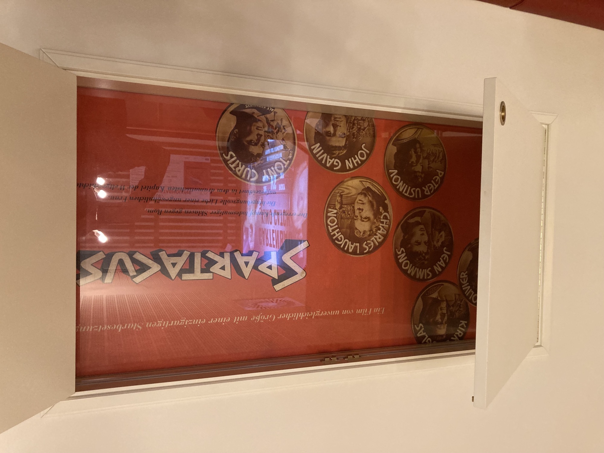hinter einer geöffneten Tür zeigt sich hinter einer Glasscheibe ein Plakat zum Film Spartacus, auf rotem Hintergrund ist in weißer Schrift mit scharf abgegrenztem Schlagschatten der Filmtitel zu lesen, die Mitwirkenden sind mit Portraits und Namen in runden Ausschnitten abgebildet, die wie Münzen wirken