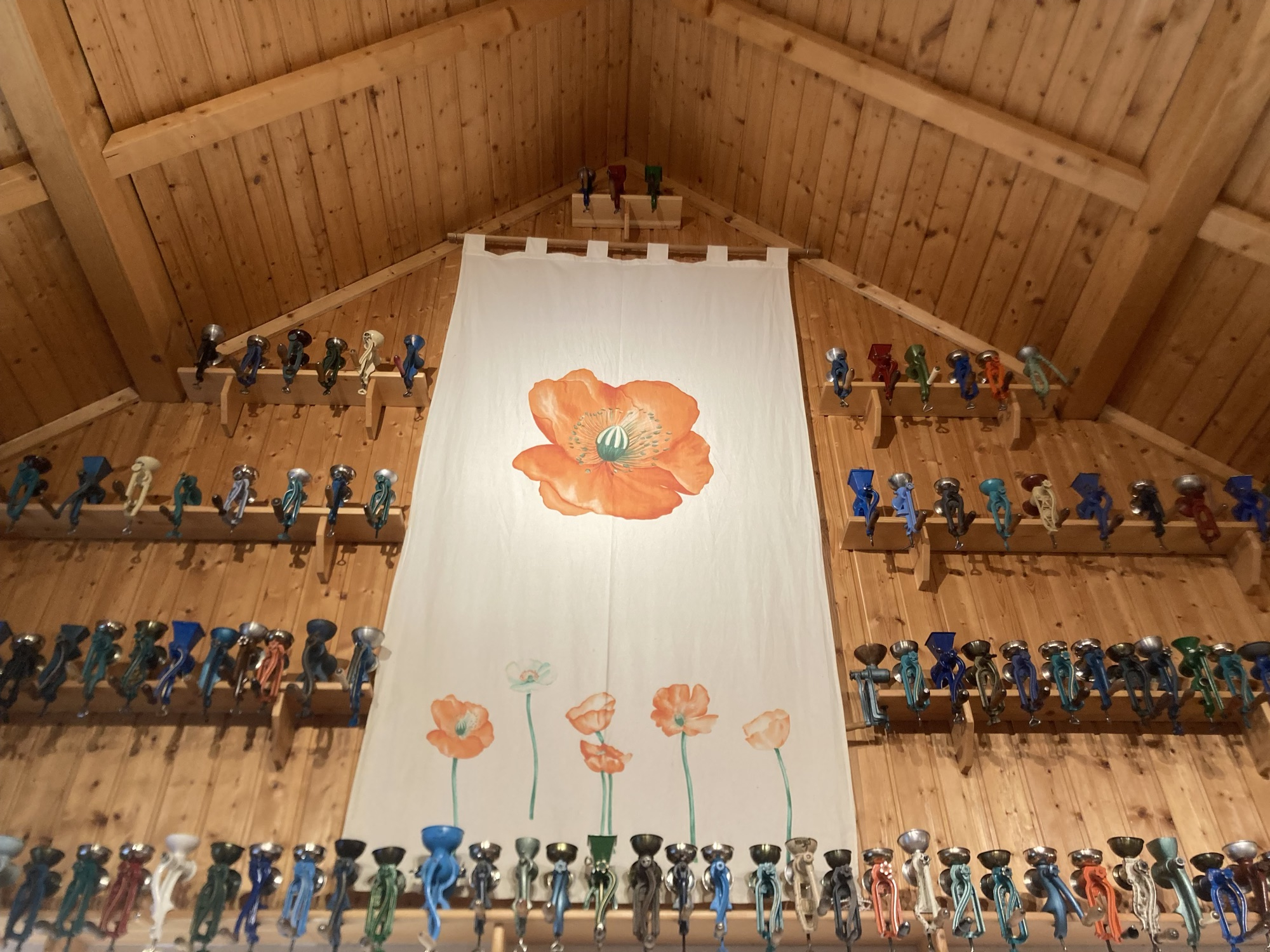 an einer Wand unter einem Spitzdach hängt ein hochformatiges Banner mit gezeichneten Mohnblumen, auf vier Regalen sind unzählige Mohnmühlen befestigt
