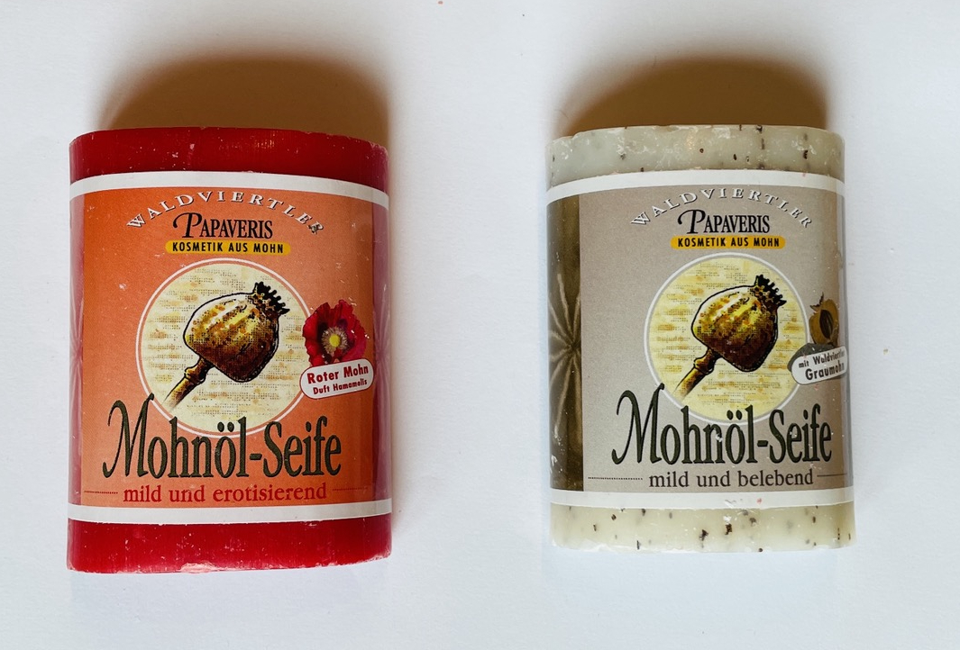 zwei Stücke Mohnölseife auf weißem Hintergrund, links eine rote Seife mit der Aufschrift „Mohnöl-Seife – mild und erotisierend“, rechts eine weiße Seife mit schwarzen Punkten mit der Aufschrift „Mohnöl-Seife – mild und belebend“