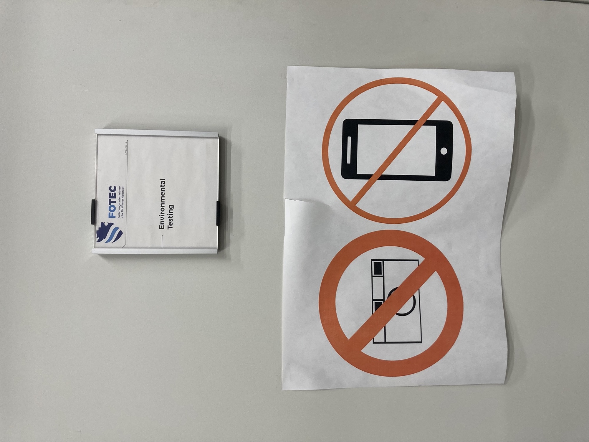 Detailaufnahme von einem Türschild, oben das FOTEC-Logo, darunter steht „Environmental Testing“, unter dem Schild ist ein ausgedruckter Zettel mit durchgestrichenem Fotokamera- und Handysymbol