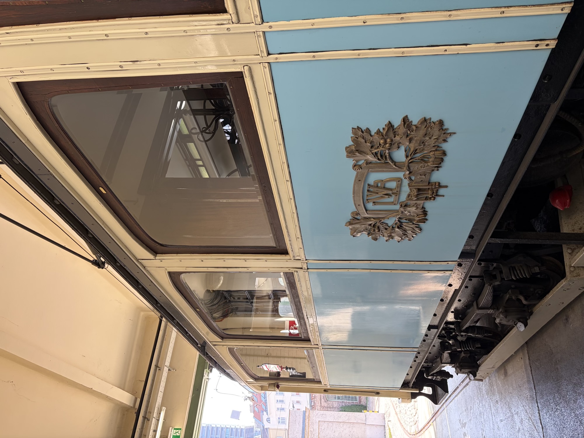 Seitenansicht eines Waggons, der unten blau und oben cremefarbig gestrichen ist, zentral im Bild ist ein Metallwappen, ein Rahmen ist von Blättern umgeben, im Rahmen die Buchstaben WLB