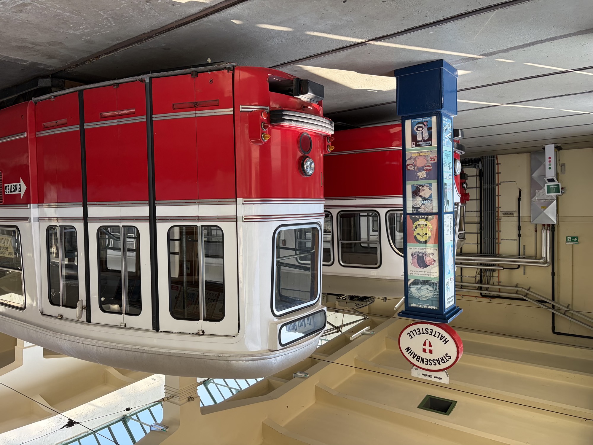 im Eingangsbereich der Remise steht ein Straßenbahnwagen mit drei Türen eben einer Straßenbahn-Haltestellen-Säule, die blaue Säule enthält diverse Werbemotive, oben darauf das Haltestellen-Symbol mit der Aufschrift „Alser Straße“