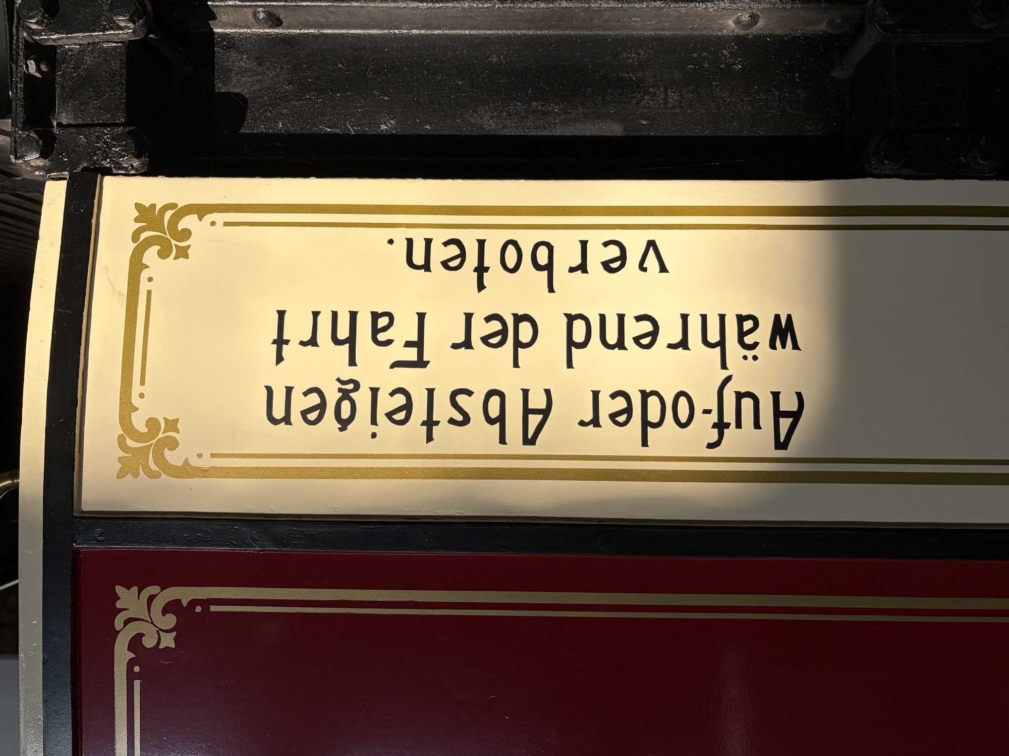 der Schriftzug „Auf-oder Absteigen während der Fahrt verboten“, schwarze Schrift auf cremefarbenem Grund in goldenem Rand, der Teil des Bildes um den Schriftzug herum liegt im Schatten
